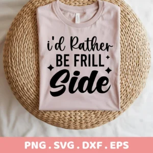 A folded light pink t-shirt with the text "I'd Rather Be Frill Side" in black, displayed on a round woven mat, representing a Frill Side SVG Design available as a digital download bundle including SVG and PNG files.